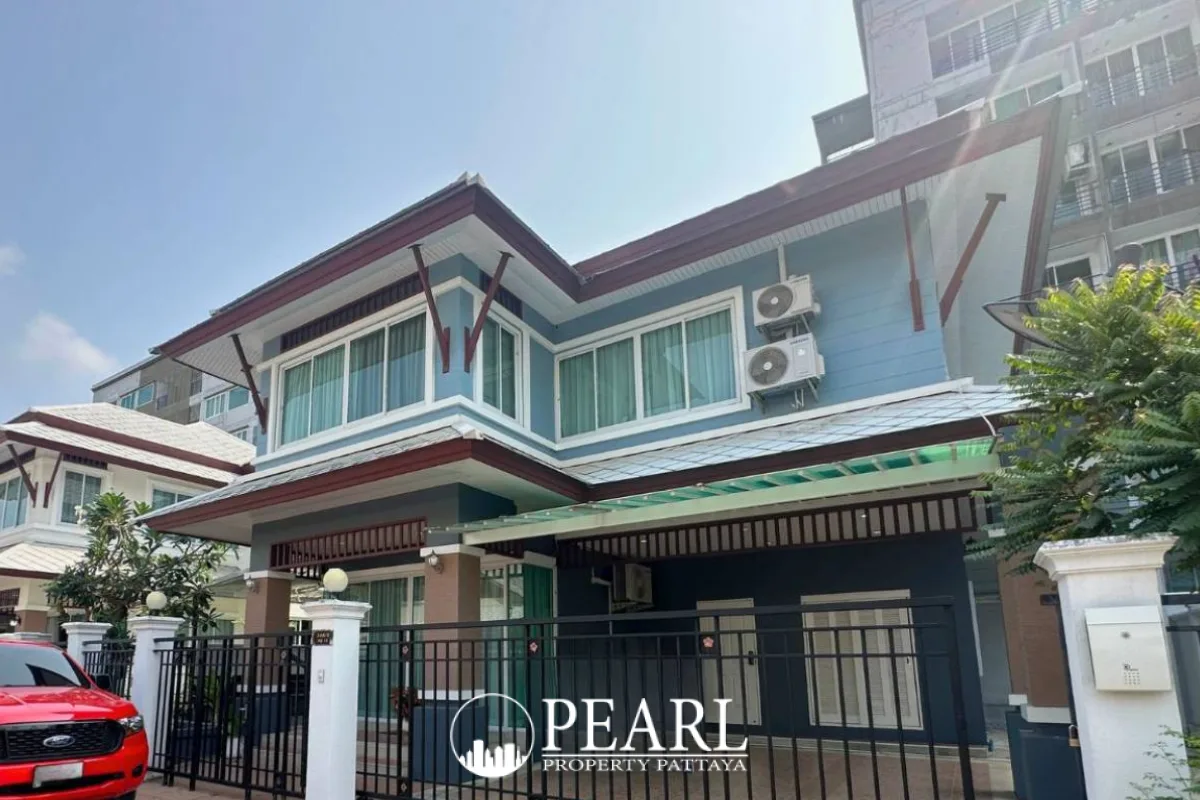 The Villa Rachawadee gated community road with white buildings, lush greenery, and a car in the distance.