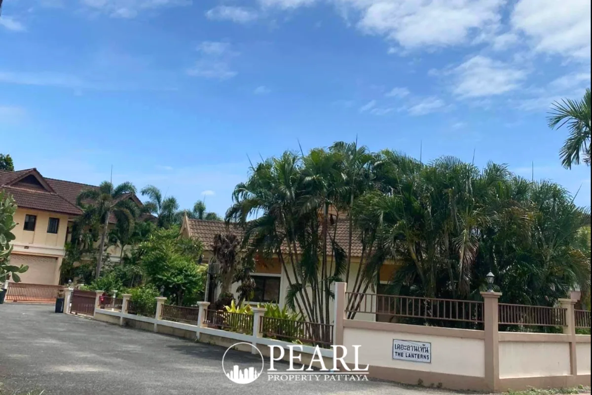 The Lanterns entrance sign on a low wall with houses and palm trees under a blue sky in Pattaya.