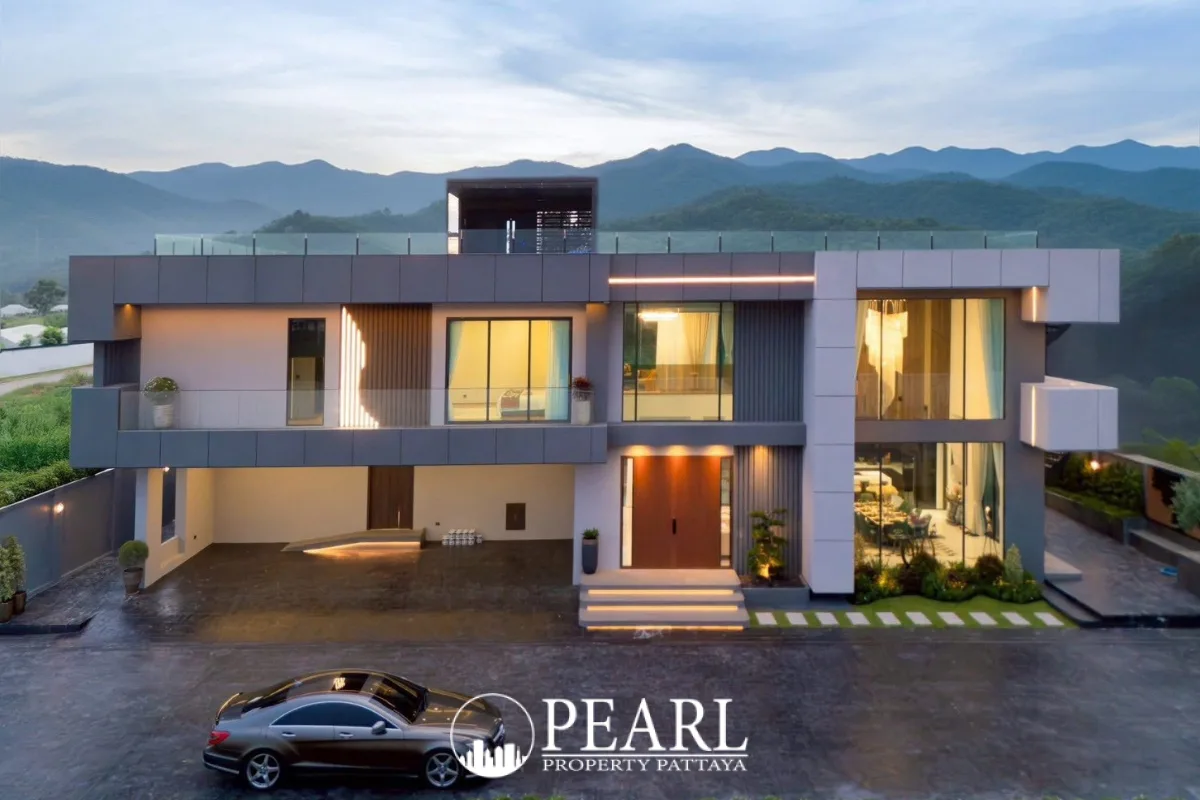 Opal Village modern house exterior with illuminated windows, glass balconies, a parked car, and mountains in the background at dusk.